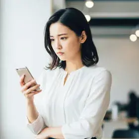 スマートフォンを手に持ち、不安げに画面を見つめる白いブラウスの女性