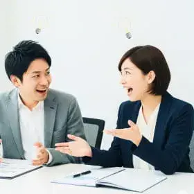 会議テーブルを挟んで向かい合い、笑顔で資料を指し示しながら話し合うビジネスパーソンの男女