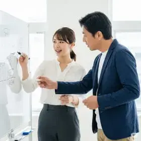 白いホワイトボードの前で、ペンを持つ女性が説明し、男性が指し示しながら話し合うビジネスシーン