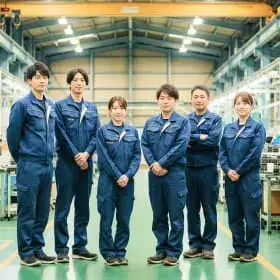 工場の作業場で青い作業着を着た6名のチームが整列し、カメラに向かって微笑む